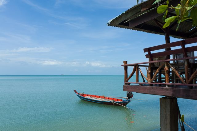 Shows longtail boat anchored near a wooden pier on a calm tropical ocean. Ideal for depicting themes of relaxation, coastal travel, tropical vacations, and serene natural scenes. Use in travel blogs, tourism promotions, beach resort advertisements, and nature-themed content.