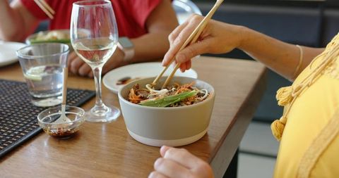 Female friends sharing noodle bowl with chopsticks at wood table, wine glass and smartwatch