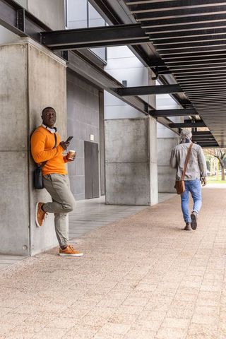 African american man leaning under modern urban walkway holding coffee and smartphone