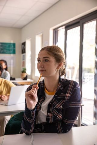 Thoughtful female student holding pencil near notebook in sunlit classroom