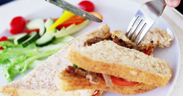 A person is cutting into a sandwich filled with vegetables and meat, with copy space. Fresh salad accompanies the meal, emphasizing a focus on wholesome, balanced eating.