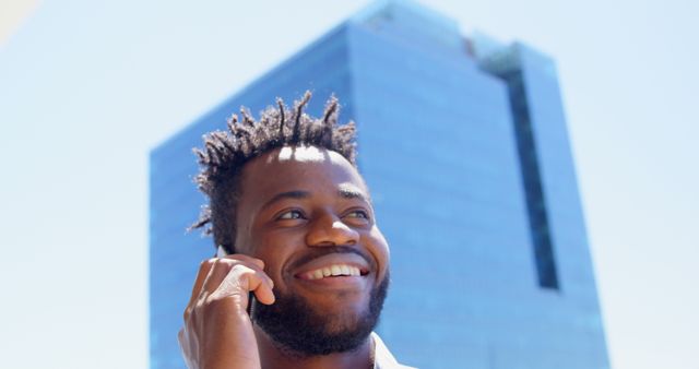 This image shows a man smiling while talking on the phone outdoors against the backdrop of a modern city building. Excellent for use in business communication themes, urban living, professional lifestyle stories, and promoting mobile technology.