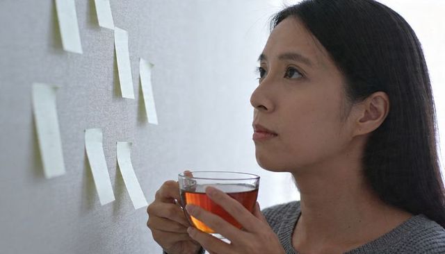 Contemplative asian woman holding tea cup while studying sticky notes on pale textured wall