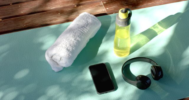 Outdoor image showing fitness gear on a turquoise yoga mat including a water bottle, white towel, black wireless headphones, and a smartphone. Ideal for promoting healthy lifestyle, summer workout routines, fitness blog posts, and wellness advertisements.