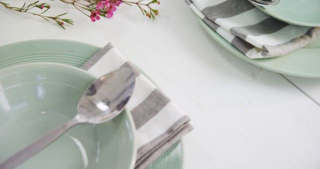 This image shows a minimalistic and elegant table layout featuring mint green dinnerware and striped napkins. The addition of a metal spoon and delicate flowers gives the presentation a touch of simplicity and grace. Ideal for articles, blog posts, or marketing materials related to home decor, table setting tips, dining etiquette, restaurant advertising, or lifestyle content.
