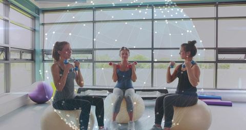 Women exercising on stability balls holding dumbbells in sunlit gym with network mesh