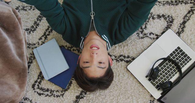 Young man lying on a cozy rug surrounded by technology items including a laptop, tablet, and headphones. Perfect for depicting relaxation, modern lifestyle, or technology use at home. Ideal for articles or advertisements about home relaxation, gadget enthusiasts, or the balanced integration of technology into daily life.