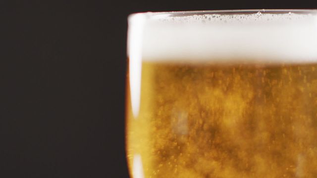 Glass of cold, bubbly beer with foam at the top sits against a moody black background, highlighting the golden hue and vibrant bubbles. Perfect for advertising related to bars, parties, alcohol brands, or social events, conveying a message of refreshment and enjoyment.