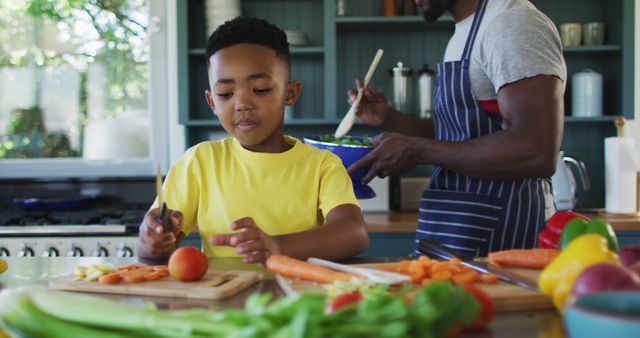 A young boy in a yellow shirt helps his father chop vegetables in a cozy kitchen. They are smiling and enjoying the cooking process together. Fresh vegetables like tomatoes, carrots, and bell peppers are visible on the counter. Great for themes related to family bonding, healthy eating, and home cooking. Perfect for use in articles, blogs, or advertisements promoting family time, nutrition, and culinary skills.