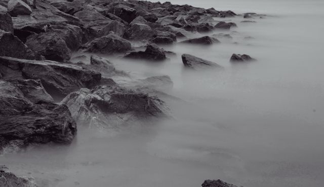 Smooth, misty water washes over large rocks at the seashore, creating a tranquil and serene coastal landscape. Monochromatic tones enhance the peaceful mood. Ideal for backgrounds, relaxation themes, nature presentations, and environmental projects.