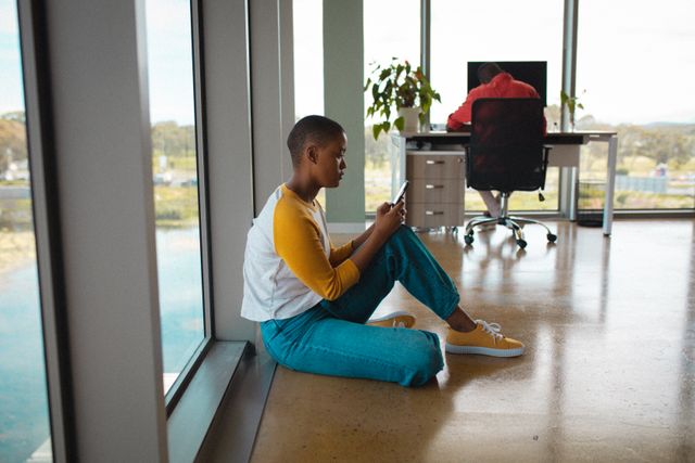 African american businesswoman in smart casuals using smart phone while sitting on floor in office. creative business, wireless technology and office workplace.