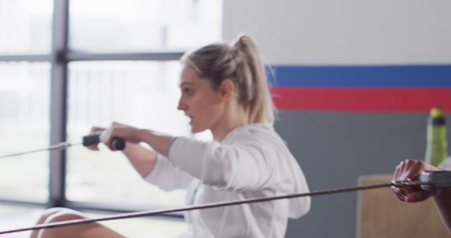 Side view of a determined Caucasian woman working out on a rowing machine in a gym. She is focused on her exercise, emphasizing strength and fitness. Ideal for use in advertising fitness programs, gym memberships, workout equipment, and healthy lifestyle promotions.