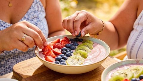 Two women arranging blueberries on pink smoothie bowl outdoors, vibrant summer brunch scene