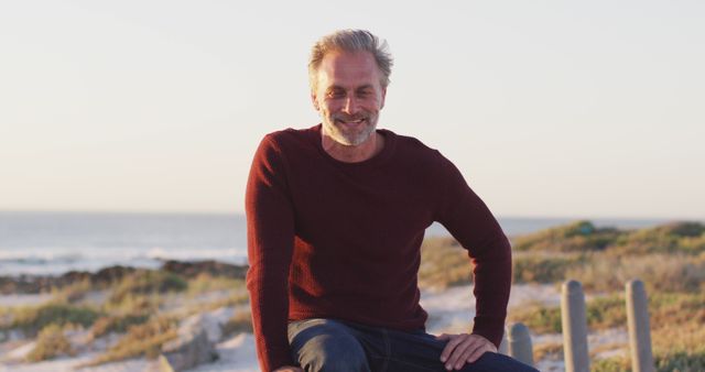 Mature man smiling and relaxing on beach at sunset, wearing casual clothing. Suitable for articles about lifestyle, travel, well-being, retirement, and outdoor activities.