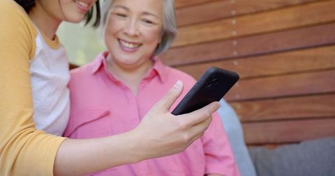 Asian mother and adult daughter smiling and sharing smartphone on sunny porch bench