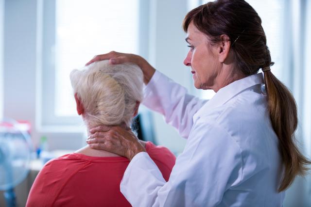 Female doctor examining senior female patient in hospital. The doctor is wearing a white coat and is gently placing her hand on the patient's head and neck. This can be used to illustrate healthcare, medical examinations, elderly care, patient-doctor relationship, or hospital settings.