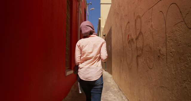 This image depicts a single woman walking through a narrow alley, framed on the left by a bright red wall and on the right by a beige wall with graffiti. The bright sunlight and clear sky indicate a warm day. This photo can be used in contexts showcasing urban exploration, solitude, adventure, and the beauty of mundane cityscapes. It can also serve as a background for content related to street photography and urban lifestyle.