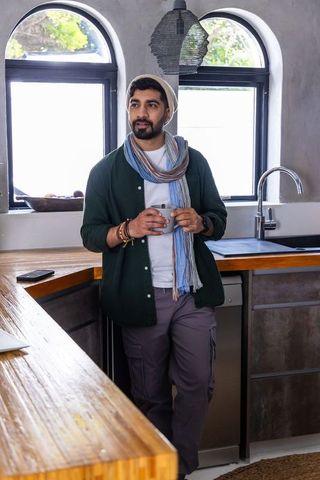 Man leaning on wood countertop holding ceramic mug, wearing scarf and beanie in cozy kitchen