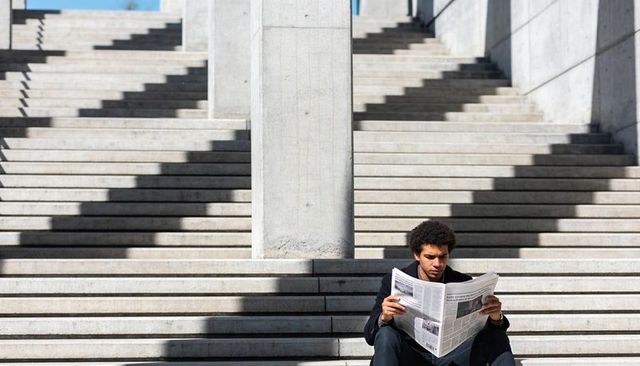 Man reading newspaper on sunlit concrete stairs with bold geometric shadow patterns