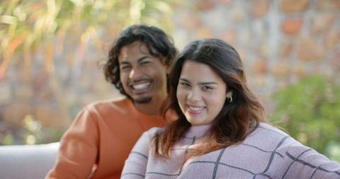Smiling couple relaxing on sunlit garden sofa, cozy moment with warm foliage and sweaters