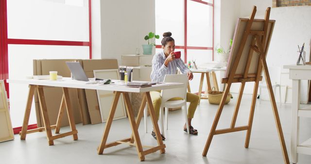 Female artist enjoying a coffee break in a bright, modern studio. This is useful for creative lifestyle blogs, encouraging relaxation, work-life balance, or illustrating artistic work environments.