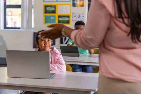 Teacher guiding asian student using laptop while classmates working on tablets