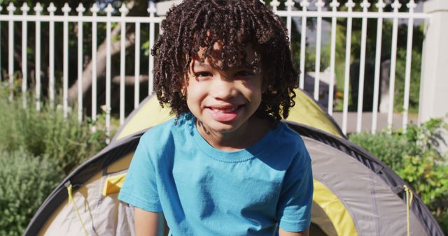 Child with curly hair sitting in front of tent in outdoor setting, smiling joyfully. Bright sunlight and garden fence in background, suggesting fun and relaxation. Useful for websites, advertisements or articles about camping, outdoor activities, children's happiness, family time, summer fun.