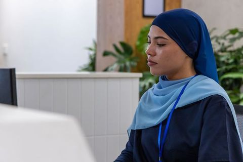 Female receptionist wearing blue hijab sitting at modern office desk focused on computer