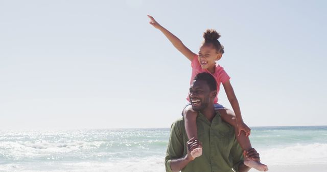 Father is carrying his young daughter on his shoulders as they enjoy a sunny day at the beach. The daughter is pointing excitedly towards the ocean. This photograph is perfect for advertisements or campaigns related to family time, vacations, father-daughter bonds, and outdoor activities. It captures happiness, interaction, and the beauty of nature, making it ideal for travel brochures, family services promotions, and lifestyle blogs.