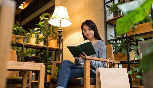 Reading woman enjoying book in cozy cafe lounge with plants and warm lamp light