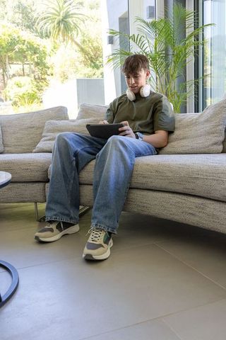 Teen relaxing on modern sectional with tablet and headphones near sunlit glass doors