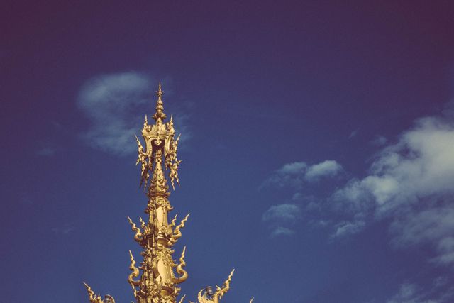 An intricately designed golden temple spire reaches towards a clear blue sky. This ornate architectural structure showcases elements of Thai culture and spirituality. Ideal for use in travel brochures, cultural exhibitions, or background for websites focusing on Asian heritage or religious tourism.