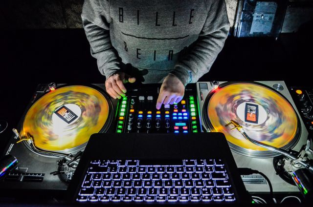 DJ operating turntables and sound mixer with colorful vinyl and illuminated laptop keyboard. Ideal for promoting nightlife and entertainment venues, DJ events, electronic music festivals, and party atmospheres.