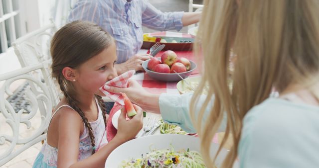 Mother wiping young daughter's face with napkin while eating salad outdoors, showing family interaction. Ideal for advertisements on family life, parenting tips, summertime activities or outdoor gatherings, and articles on healthy eating or family bonding moments.