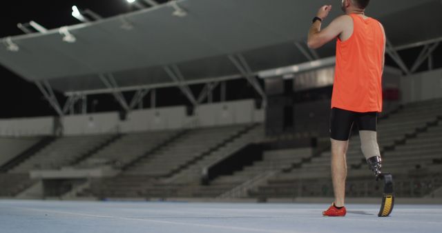 Athlete with prosthetic leg running on a track at night in a stadium. The focus is on the back of the athlete who is wearing a bright orange sports top and black shorts. This image can be used to illustrate themes of determination, fitness, sports, and overcoming challenges. It is suitable for use in articles, blogs, promotional materials, and advertisements related to sports, adaptive athletes, and motivation.