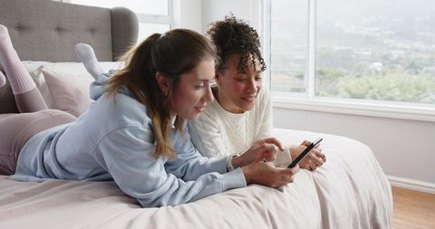 Two friends sharing smartphone on bed in sunlit pastel bedroom with hillside view