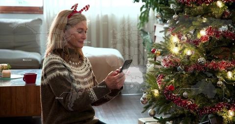 Woman using smartphone by christmas tree wearing knit sweater and candy-cane headband