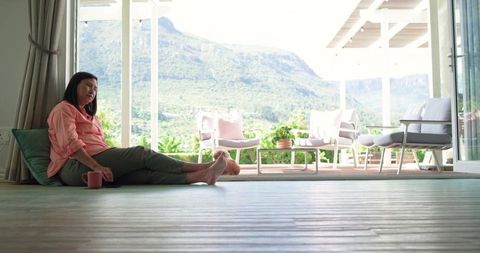 Senior woman sitting on floor by sliding doors, relaxing with mug, mountain terrace view