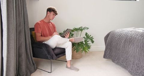 Young man sitting in modern bedroom armchair using tablet for remote work and relaxation