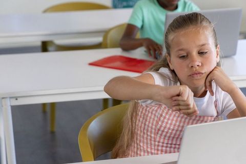Schoolgirl checking wrist while using laptop, classmates learning in modern diverse classroom