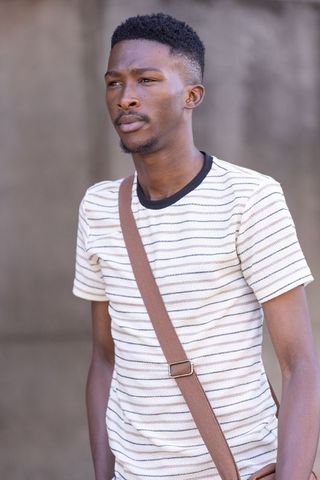 Contemplative young man standing wearing cream striped tee and brown crossbody strap