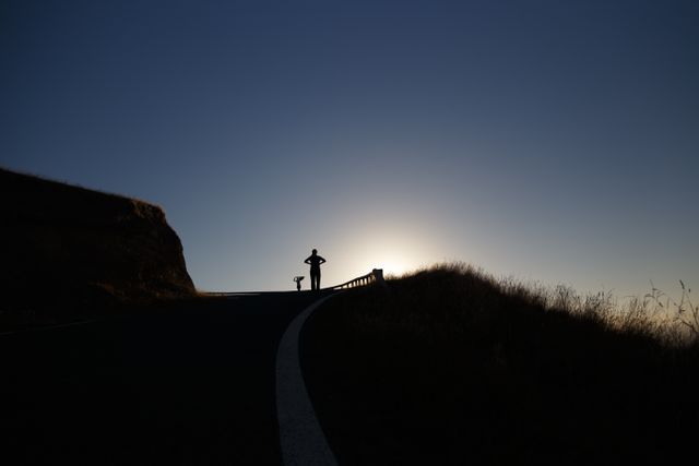 Person standing on roadside facing sunset. Can be used for themes of adventure, travel, solitude, and nature. Ideal for motivational content, travel blogs, or backgrounds portraying tranquility and natural beauty.