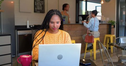 Young woman typing on laptop in cozy cafe wearing yellow knit sweater with pink mug and barista coun