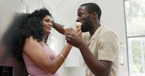 Diverse couple sharing ice cream cones, smiling and laughing in sunlit modern kitchen