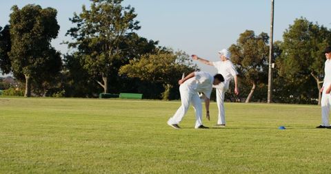 Diverse male cricketers practicing bowling and fielding on sunlit grass with blue cone