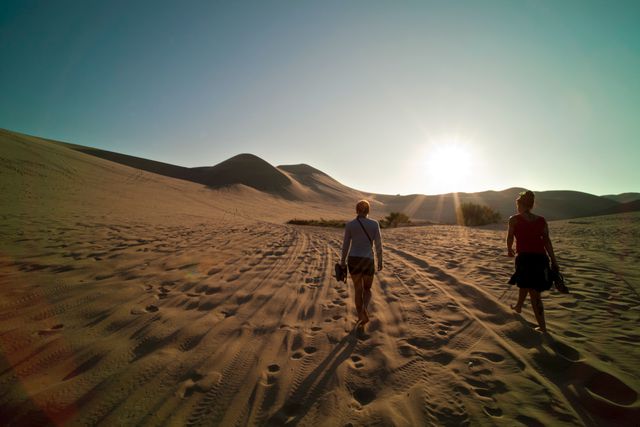 Two women hiking through desert dunes at sunset, casting long shadows on the sandy terrain. Great for travel blogs, adventure magazines, landscape collections, nature exploration themes, and outdoor lifestyle promotions.