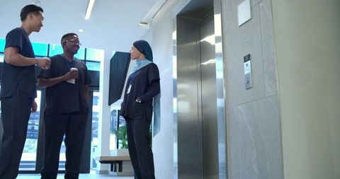 Multicultural healthcare team chatting in modern hospital lobby by elevator with coffee