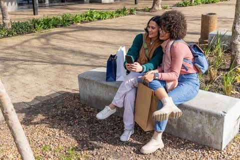 Friends taking selfie, carrying shopping bags, sitting on sunny urban bench