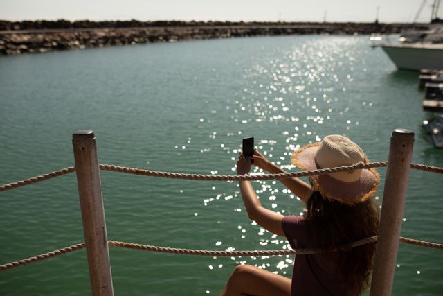 Rear view of teenage Caucasian girl, wearing a straw hat, enjoying her time on a promenade by the sea, on a sunny day, sitting and taking photos on her smartphone.