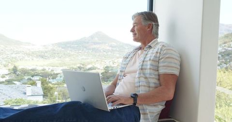 Senior man working remotely on laptop at window, gazing over sunlit valley landscape